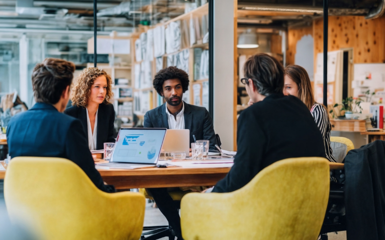 Group of professionals seated around a conference table, engaged in discussion while using laptops.