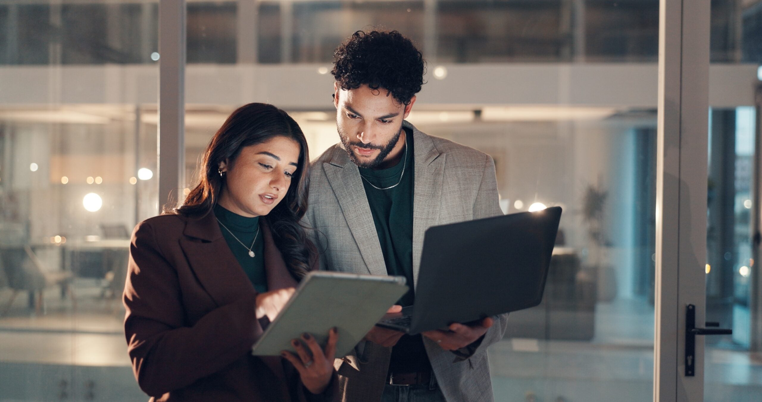 A man in a modern office holding a laptop while looking at a woman’s tablet screen during a discussion.
