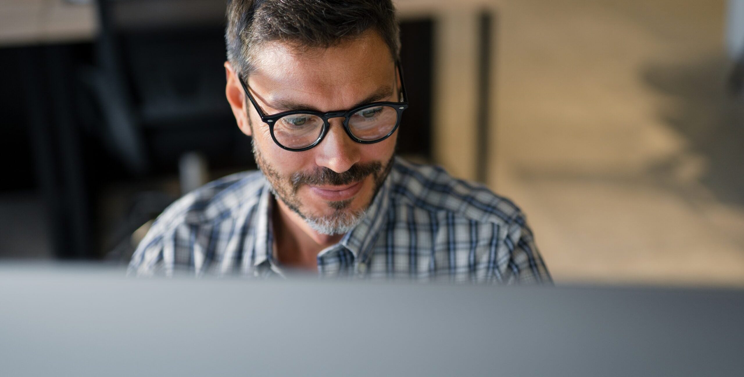 Man wearing glasses works on a computer at home, with a monitor screen reflected in his lenses.